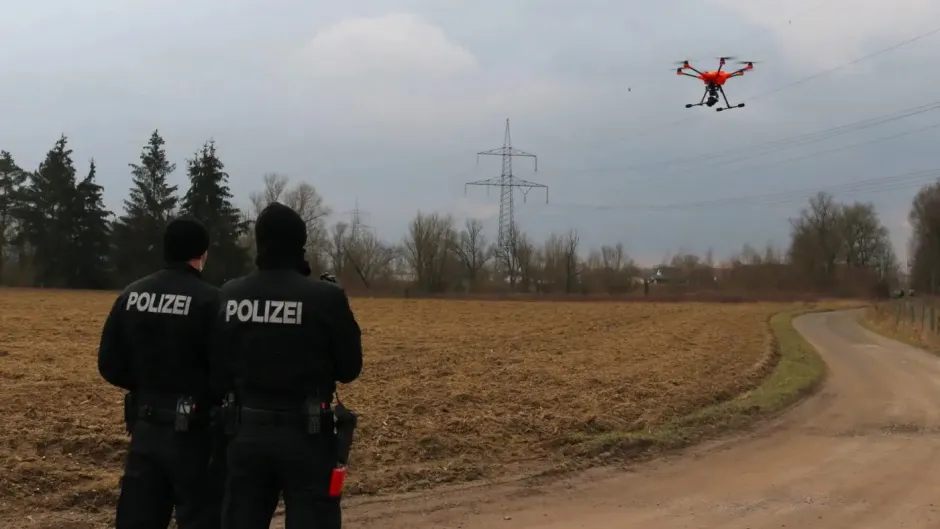 Zwei Polizist*innen stehen vor einem gepflügten Feld, vor ihnen fliegt ein Multicopter/Drohne.